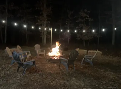 Fire pit burning at night under strung festoon lighting, ringed by chairs